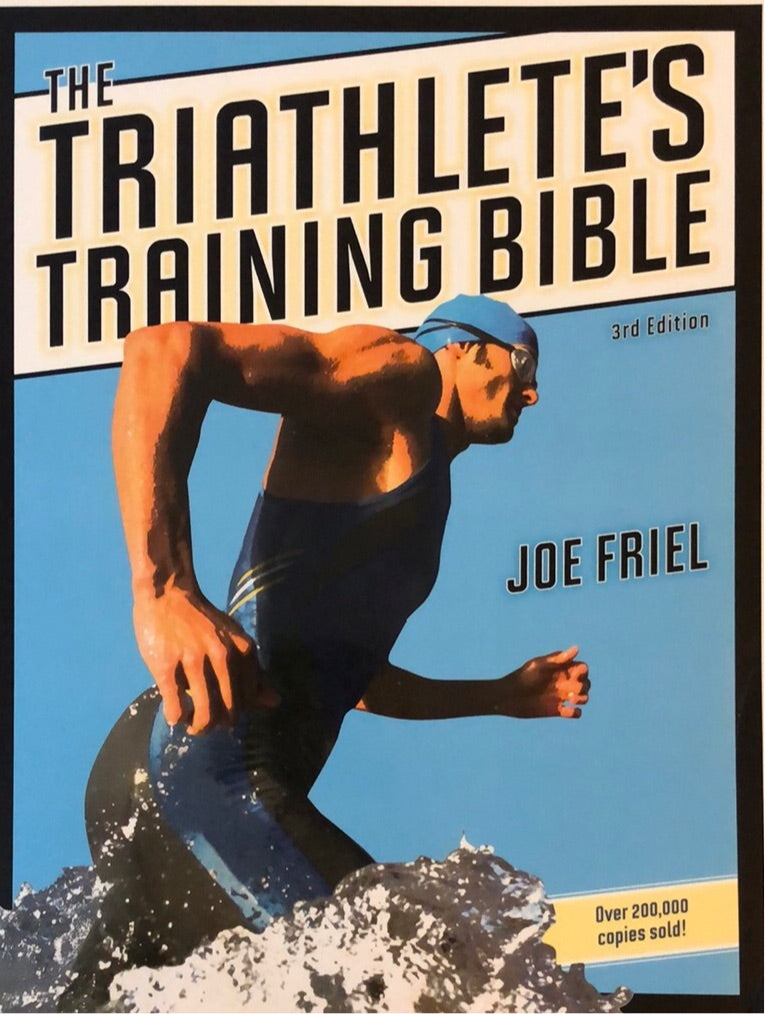 Bokillustrasjon av "The Triathlete's Training Bible" i pocketutgave, med åpen bok og treningsplaner på sidene